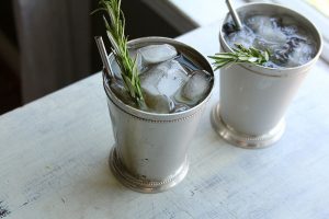 2 ice filled Little Britches cocktails in metal cups with rosemary and a straw.
