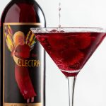 Electra Berry cocktail made with Vodka bring poured in a martini glass with raspberries and bottle of Red Electra Moscato behind it.