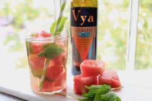 Watermelon mojito cocktail with mint and watermelon garnishes next to a bottle of Vya Whisper Dry Vermouth in front of a window.