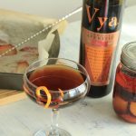 Plum Roasted Manhattan cocktail in a coupe glass and a mason jar next to a bottle of Vya Sweet Vermouth and a baking pan filled with roast plums.