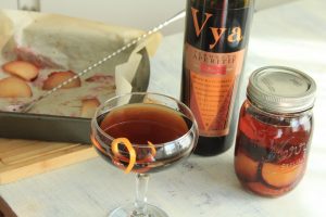 Plum Roasted Manhattan cocktail in a coupe glass and a mason jar next to a bottle of Vya Sweet Vermouth and a baking pan filled with roast plums.