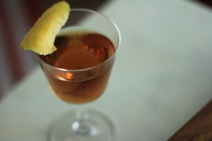 50 50 Manhattan cocktail in a nick and nora glass with a lemon peel garnish.