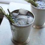 2 ice filled Little Britches cocktails in metal cups with rosemary and a straw.