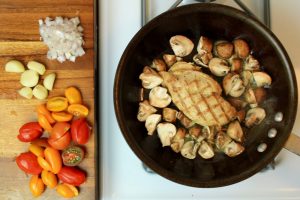 Grilled chicken breast and mushrooms in a pan next to a cutting board filled with cherry tomatoes, garlic and chopped onion.