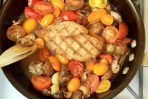 Grilled chicken breast on top of tomatoes, mushrooms, onions and garlic in a pan.