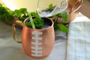 Cocktail being pouring into a football shaped mule mug with basil, ice and straw.