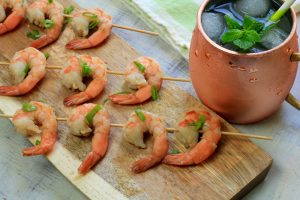 Cooked shrimp on skewers with green onion and a cocktail filled football shaped mule mug in the background.