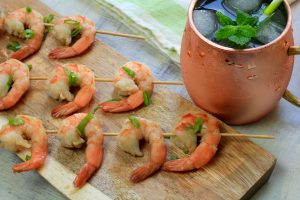 Cooked shrimp on skewers with green onion and a cocktail filled football shaped mule mug in the background.