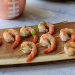 Cooked shrimp on skewers with green onion and a football shaped cup in the background.