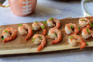 Cooked shrimp on skewers with green onion and a football shaped cup in the background.