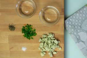 Pepper, blue cheese crumbles and green onions next to two bowls on a cutting board.