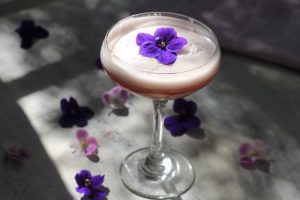 Low proof cocktail in a coupe glass with an edible flower on top and more edible flowers around it.