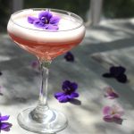 Low proof cocktail in a coupe glass with an edible flower on top and more edible flowers around it in front of a window.