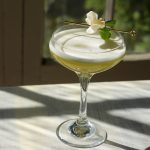 Spring cocktail in a coupe glass with an edible flower garnish in front of a window.