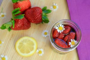 Strawberries being infused in Vya Sweet Vermouth and Deviation dessert wine in a mason jar next to lemon, strawberries and edible flowers.