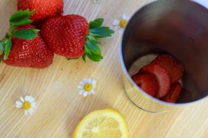 Sliced strawberries in a metal cup with strawberries, edible flowers and a lemon next to it.