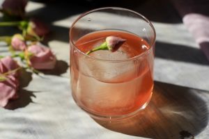 Low proof cocktail in a cocktail glass with a large ice cube and edible flower on top next to pink roses.