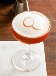 Negroni flip cocktail in a coupe glass with a Q written in the egg white topping.