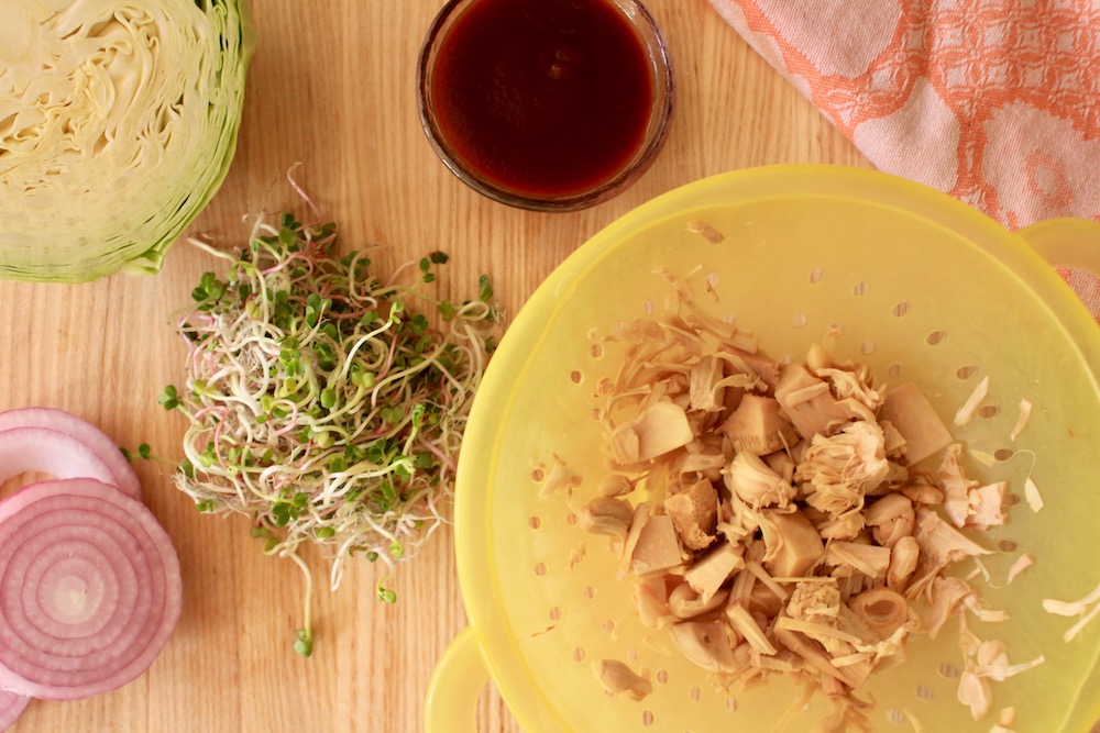 Shredded jack fruit in a bowl next to onions, sprouts and cabbage.
