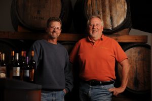 Quady winemakers Darin Peterson and Michael Blaylock standing next to each other in front of wine barrel.