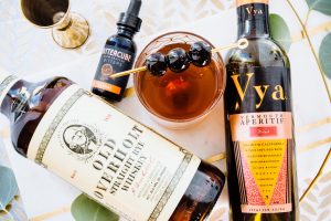 Bottle of Vya Sweet Vermouth, Bittercube bitters and Old Overholt Rye Whiskey lying on a table next to a Manhattan cocktail with maraschino cherry garnish.