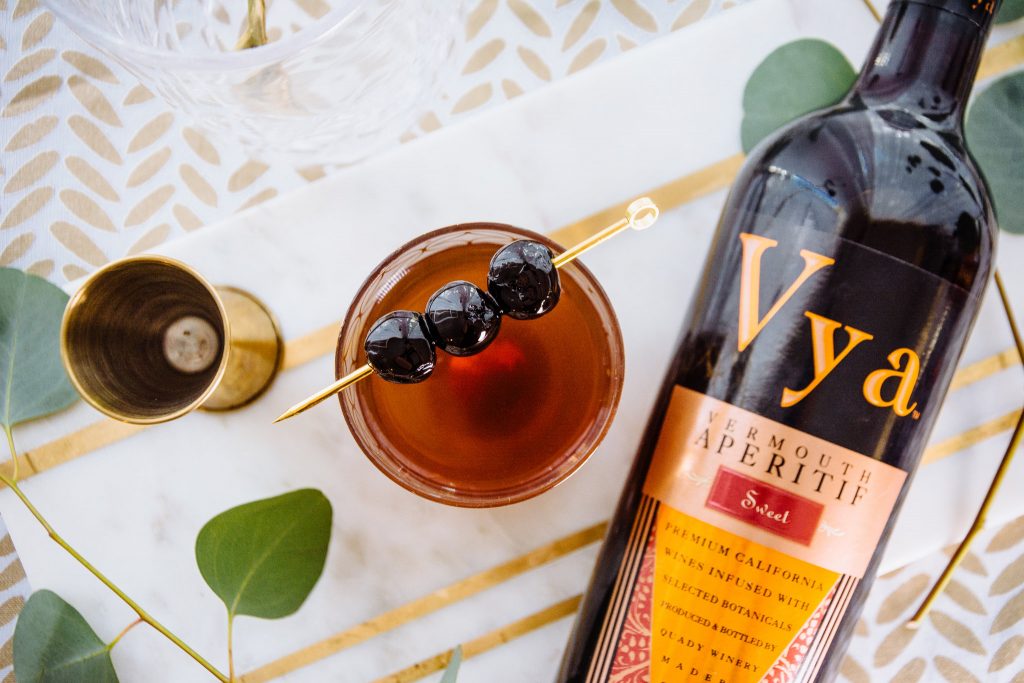 Bottle of Vya Sweet Vermouth and jigger lying on a table next to a Manhattan cocktail with maraschino cherry garnish.