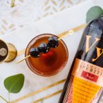 Bottle of Vya Sweet Vermouth and jigger lying on a table next to a Manhattan cocktail with maraschino cherry garnish.
