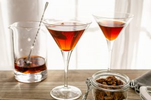 Two Pecan Manhattan cocktails in martini glasses in front of a window on a table next to pecans and cocktail stir glass and spoon.