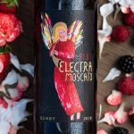 Close up of Red Electra Moscato wine bottle and label on a wood setting surrounded by flowers and berries.