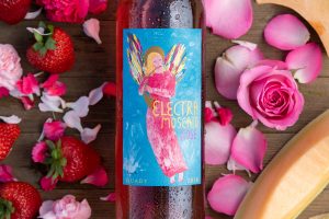 A close up of the new Electra Moscato Rosé surrounded by flowers, roses, fresh strawberries and melon.