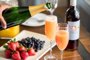 Hand pouring sparkling wine into champagne flute with Elysium black muscat dessert wine and orange juice inside, surrounded by dishes, fruit and a bottle of Elysium.