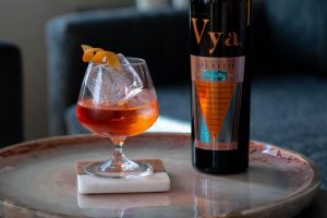 Aperol negroni cocktail created by Beautiful Booze sitting on a coffee table next to a bottle of Vya Whisper Dry Vermouth.