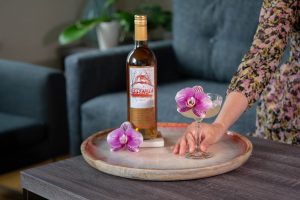 A hand holding a Margarita cocktail created by Beautiful Booze sitting next to a bottle of Quady Essensia Orange Muscat Dessert Wine and an edible orchid flower on a coffee table.