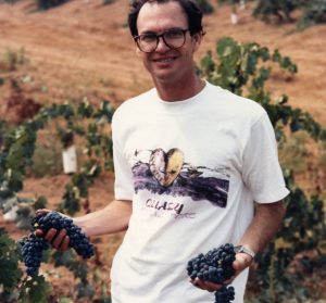 Vintage photo of Andrew Quady holding two bunches or red grapes in front of a vineyard.