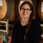 Andrea Milliorn, Senior Accountant at Quady Winery, holding a glass of Electra Moscato in front of a wall of wine barrels.