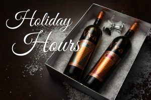 A bottle of Vya Sweet Vermouth and Vya Extra Dry Vermouth lying down in a gift box on snow with text next to them, "Holiday Hours."