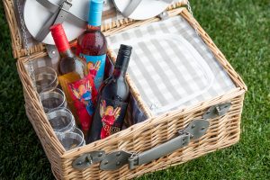Three wine bottles sitting in a picnic basket with four glasses inside. The basket is sitting on top of grass.