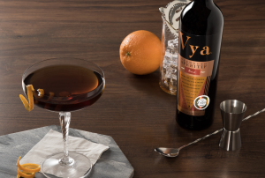 Vya bottle sitting on a table next to an orange a stirring spoon and the glass with liquid inside is a manhattan cocktail sitting on a marble tray
