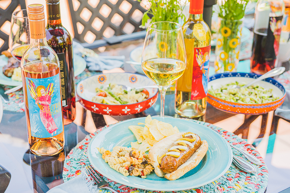 A plate with a brat, pasta salad and chips, with bottles of Electra Moscato